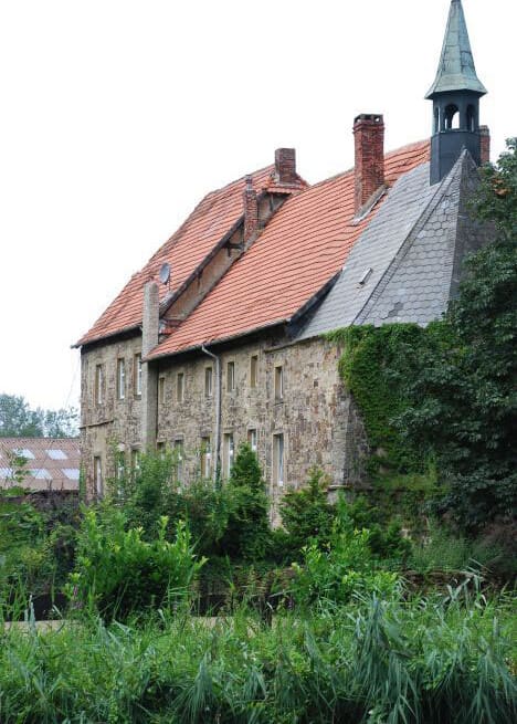 Das Gutshaus aus Bruchstein mit rotem Dach und dem Türmchen der Hauskapelle, davor hohes Grün